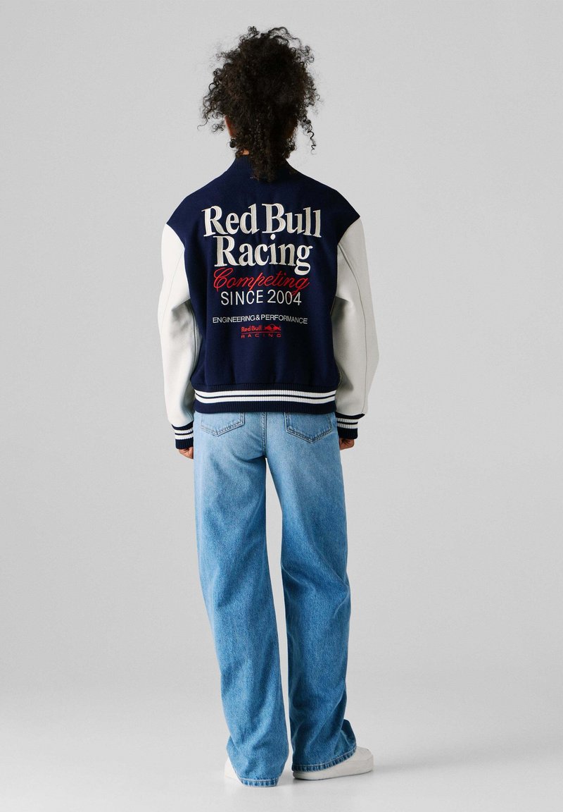 Personne aux cheveux bouclés portant une veste Red Bull Racing bleu marine et blanche, ainsi qu'un jean bleu clair, debout devant un fond gris uni.