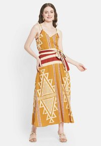 Maxikleid in Senfgelb mit cremefarbenen, gemusterten geometrischen Designs. Mit V-Ausschnitt, Rüsche-Trägern und einem roten Taillengürtel. Glatte Textur.