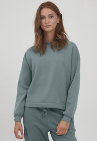 Felpa con cappuccio in morbido tessuto teal tenue. Presenta una vestibilità rilassata, polsini a costine e un cordoncino all'orlo. Texture liscia, design minimale.