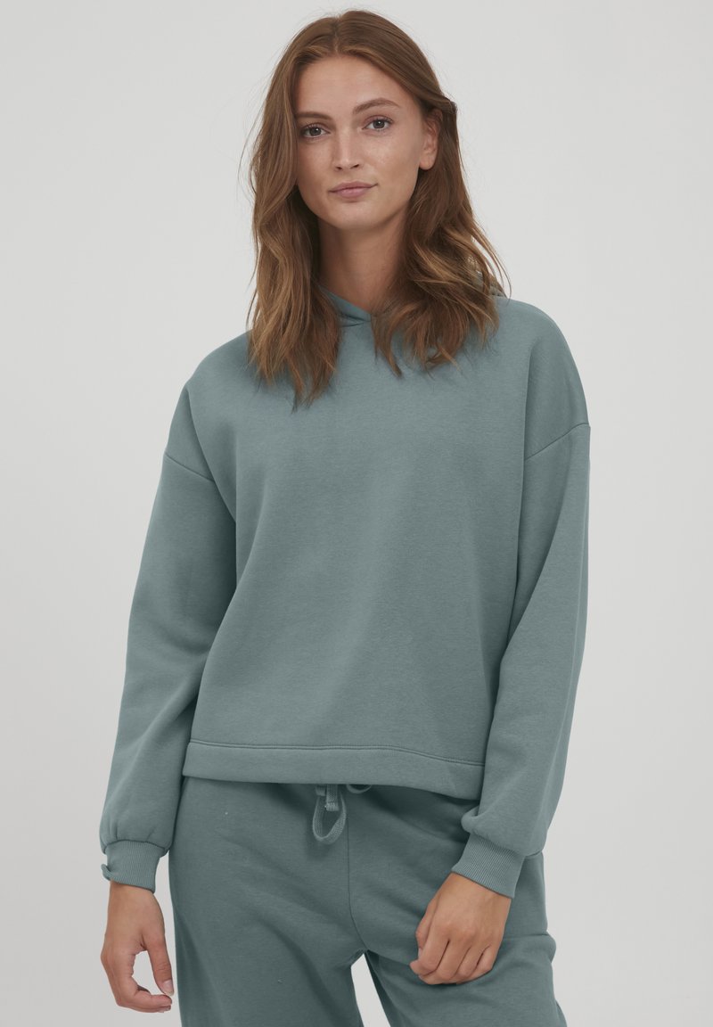 Hooded svetrovka iz mehkega, prigušenega teal blaga. Ima sproščen kroj, rebraste manšete in vrvico na robu. Gladka tekstura, minimalističen dizajn.