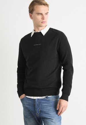 Pull en maille noir avec texture côtelée et col rond, porté sur une chemise blanche à col. Présente un logo discret sur la poitrine.