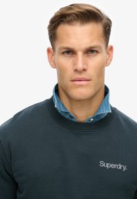 Sudadera verde oscura con cuello redondo acanalado, que presenta un logo "Superdry" en gris claro. Se lleva sobre una camisa de denim con cuello abotonado.