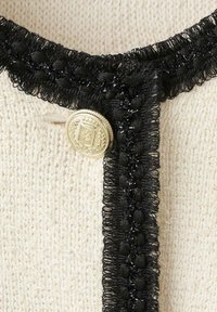 Tissu en maille crème avec bordure en dentelle noire et un seul bouton doré orné d'un blason embossé et d'un détail de couronne.