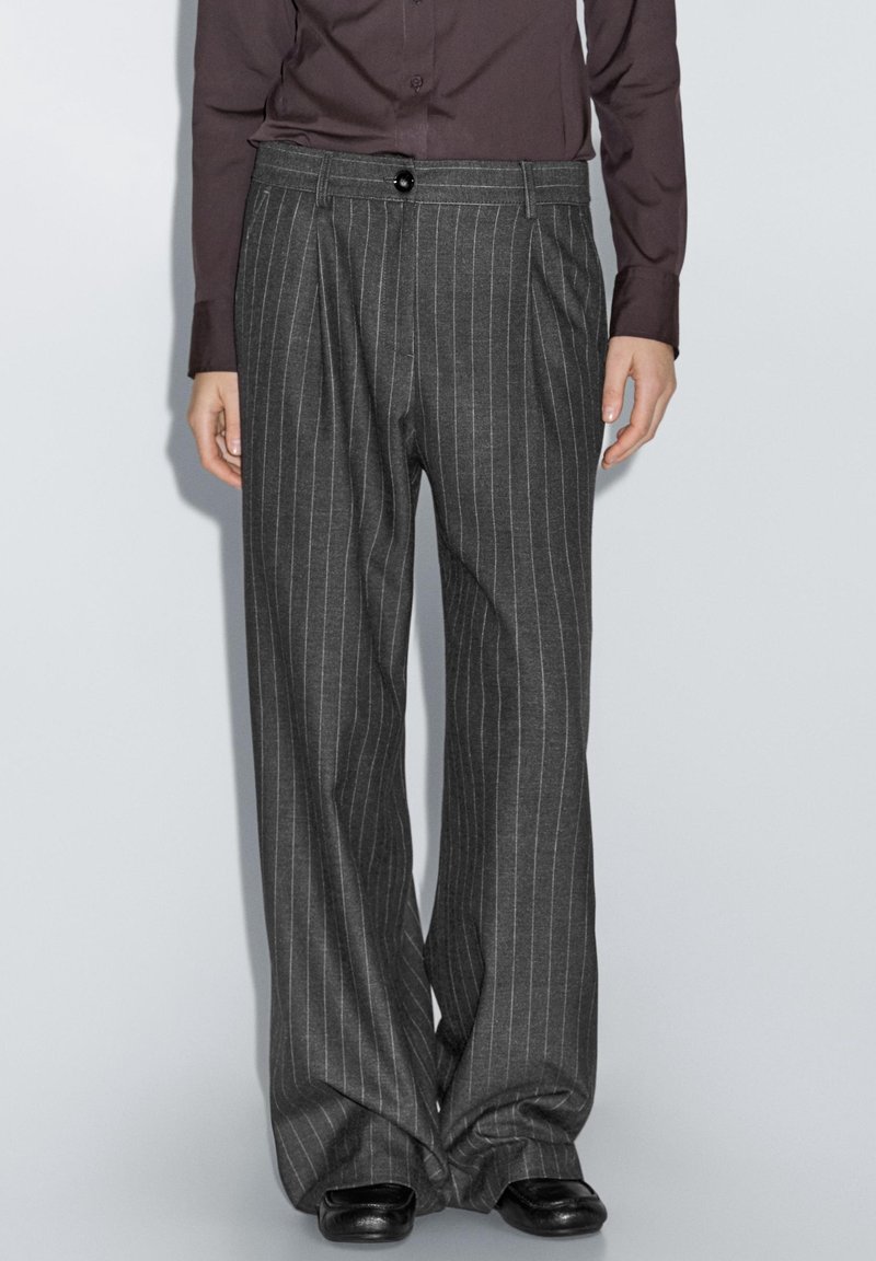 Personne portant un pantalon large gris foncé à rayures fines et des chaussures noires avec une chemise sombre à manches longues, debout devant un fond uni.