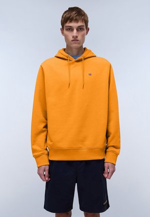 Yellow hoodie with front pockets, drawstring, ribbed cuffs, and hem. Worn over a blue shirt, paired with black shorts. Simple logo accent.