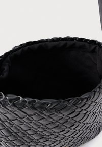 Μαύρη υφασμάτινη τσάντα με υφή, με στρογγυλεμένο άνω μέρος και άνευ φόδρας εσωτερικό. Το υλικό είναι λαμπερό και ευέλικτο.