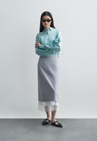 Šviesiai mėlynas marškiniai, pilkas pieštuko sijonas su nėriniu, ir juodi batai su plokščiu padu. Modelis stovi rankomis sukryžiuotomis priešais vientisą foną.