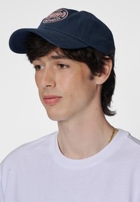 Marineblaues Cap mit einem runden Schirm und gesticktem Logo in Pink. Kombiniert mit einem schlichten weißen T-Shirt, das einen lässigen Stil zeigt.