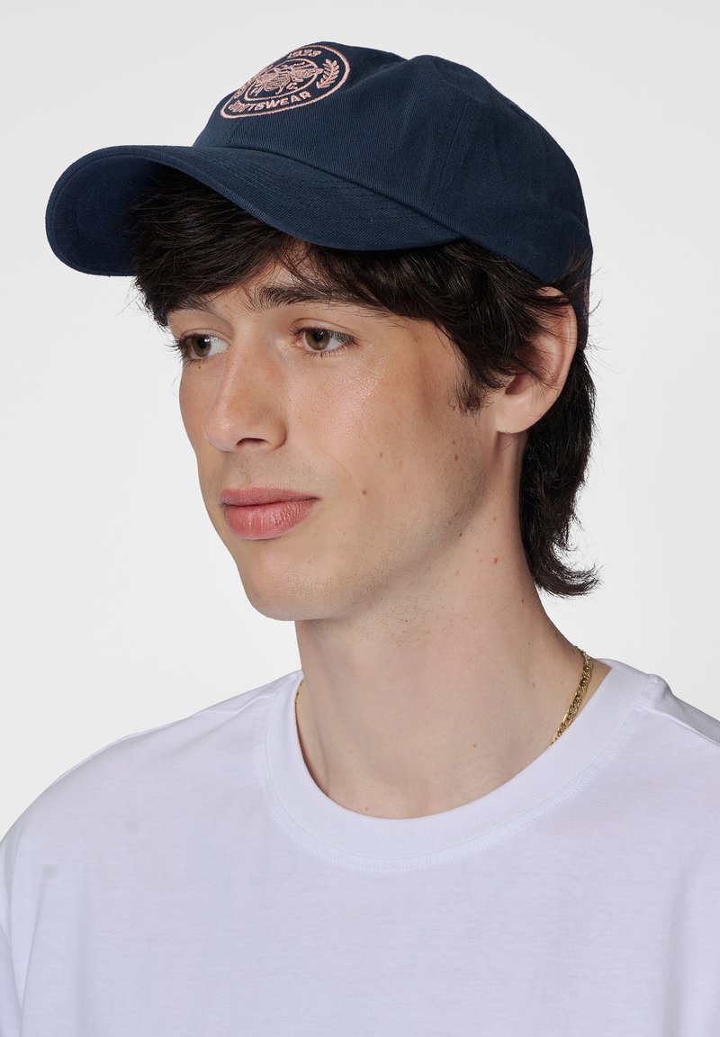 Marineblaues Cap mit einem runden Schirm und gesticktem Logo in Pink. Kombiniert mit einem schlichten weißen T-Shirt, das einen lässigen Stil zeigt.