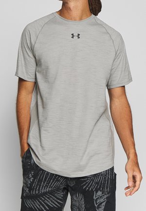 Homme portant un t-shirt à manches courtes Under Armour gris clair et un short noir à motifs, une main dans la poche, légèrement tourné vers la gauche.