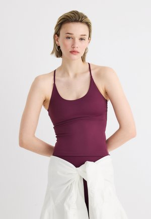 Γυναίκα που φοράει μπουστάκι χωρίς μανίκια σε μπορντό χρώμα και αντίστοιχο κάτω μέρος, με ένα λευκό πουκάμισο δεμένο γύρω από τη μέση της, με φόντο απλό.