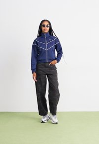 Mornarica zip-up jakna s belim obrubom, hLAče s kamuflažnim vzorcem in sive superge. Model nosi sončna očala in ima dolge kite.