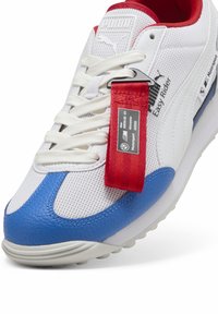 Weiße Netz- und blaue Leder-Sneaker mit roten Akzenten, ausgestattet mit einem auffälligen Etikett, dämpfender Sohle und strukturierten Details entlang des Obermaterials.