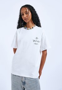 Bílé bavlněné tričko s krátkými rukávy, s grafickým potiskem na levé straně a volnějším střihem. Spárované se světle modrými džíny.