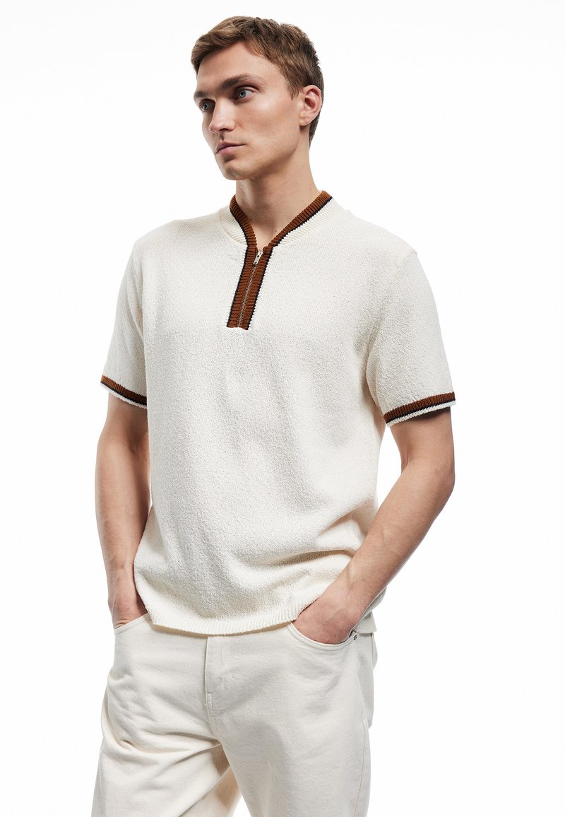 Kurzarmpullover in creme mit strukturierter Oberfläche. Verfügt über einen braunen Reißverschlusskragen und kontrastierende braune Akzente an den Ärmeln. Legere Passform.