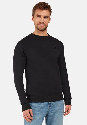 Man draagt een zwarte trui met lange mouwen en blauwe spijkerbroek, staand tegen een effen lichte achtergrond, kijkt recht vooruit.