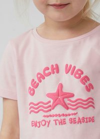 Kurzärmliges, pinkes Baumwoll-T-Shirt mit erhabenem, pinken Text "BEACH VIBES", einem Seeigel-Grafik und wellenförmigem Design. Weiche Textur, lässige Passform.