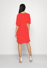 Rode jurk tot op de knie met korte pofmouwen, getailleerde taille en een vloeiende silhouet. Gecombineerd met witte, open-toe hakken.