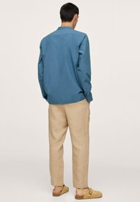 Camicia blu a maniche lunghe in tessuto leggero, pantaloni di lino beige e scarpe slip-on marroni, viste da dietro.