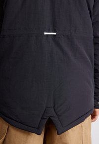 Schwarze leichte Jacke mit einem Saum, der vorne kürzer und hinten länger ist, und einem weißen Kordelzug am Rücken. Glatte Textur, minimalistisches Design und keine sichtbaren Beschläge.