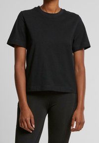 Persoon in een eenvoudig zwart T-shirt met korte mouwen en zwarte broek, staand tegen een neutrale grijze achtergrond.