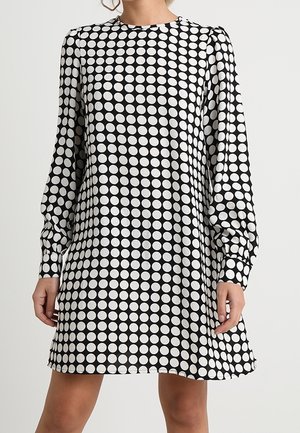 Vrouw met een zwart-witte stippenjurk met lange mouwen, een ronde hals, boven de knie en met gerimpelde manchetten.