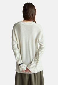 Weißer Strickpullover mit langen, gerippten Ärmeln und lockerer Passform, mit einem runden Halsausschnitt und strukturierten Details auf der Rückseite.