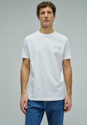 Homme portant un t-shirt blanc à manches courtes avec le texte « SALSA JEANS » sur la poitrine et un jean bleu, debout devant un fond gris uni.