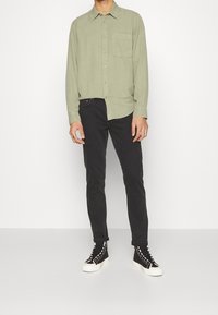 Hombre con camisa de botones de color verde claro, jeans ajustados negros y zapatillas de caña alta negras con suelas blancas, de pie frente a un fondo liso.