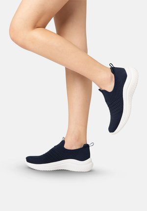 Osoba nosząca granatowe dziergane buty typu slip-on z grubymi białymi podeszwami, jedna stopa uniesiona, druga płasko na jasnoszarej podłodze.