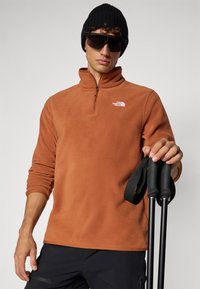 Pull-over en flanelle orange avec un col zippé mi-haut et un logo blanc. Bonnet en tricot noir et lunettes de soleil sombres. Bâtons de randonnée noirs tenus à la main.