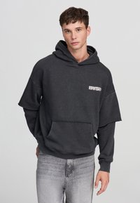 Felpa con cappuccio grigia oversize con tasca frontale e orlo grezzo. Tessuto morbido, design casual, logo bianco a contrasto sul lato sinistro del petto.