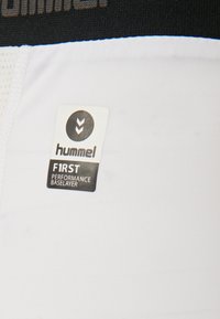 Baselayer performance bianco con cintura nera. L'etichetta riporta "hummel" e "F1RST PERFORMANCE BASELAYER" in testo nero. Tessuto morbido.