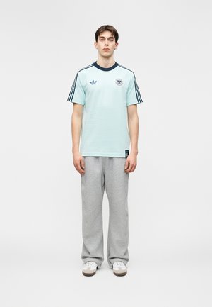 Mladi moški stoji pokončno, oblečen v svetlo modro Adidas majico s črnimi črtami in sive športne hlače na belem ozadju.