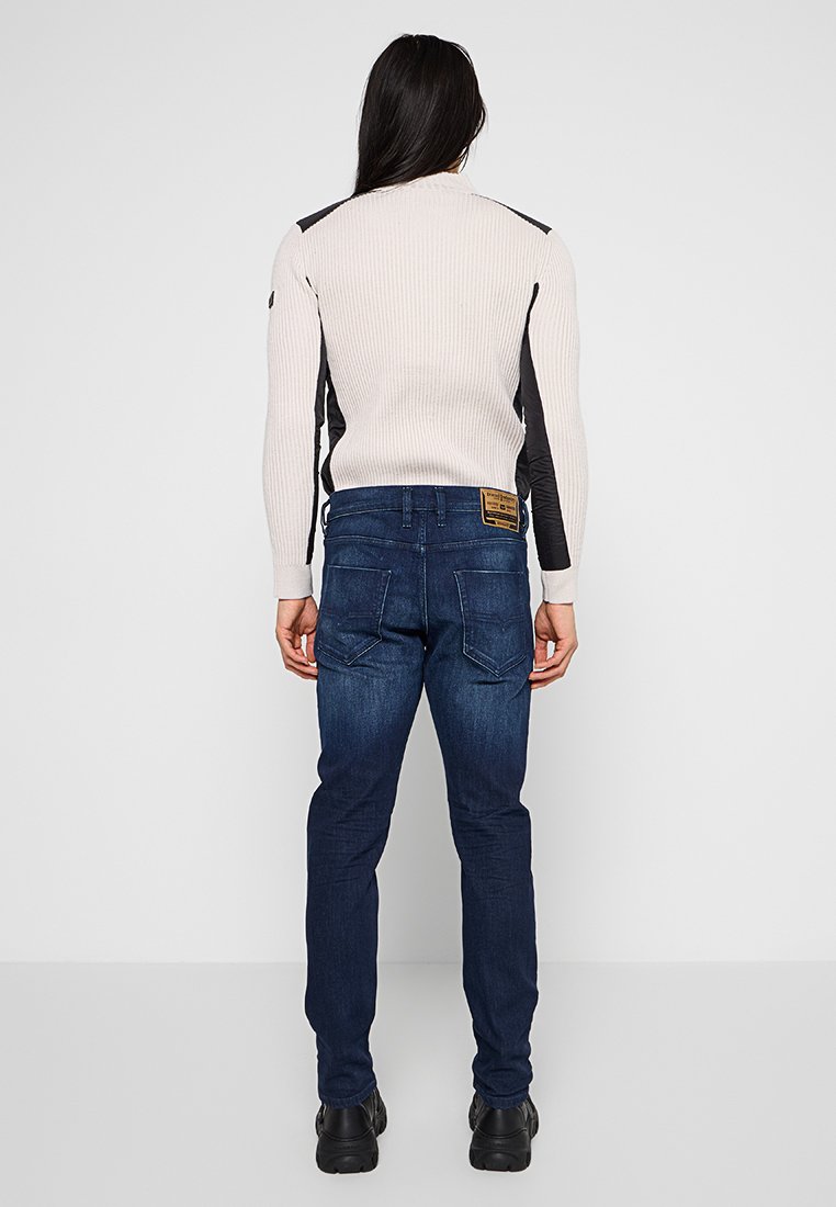 Jean en denim bleu foncé avec une coupe slim, doté de deux poches arrière et d'une pièce en cuir marron à la taille. Texture lisse et robuste.