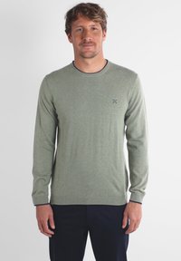 Pull lumineux vert à manches longues, col rond, avec des poignets et un bas côtelés, présentant un petit logo brodé sur la poitrine gauche.