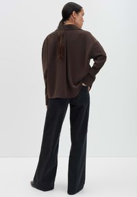 Femme aux longs cheveux foncés en queue de cheval basse, portant un jeans noir ample et une chemise marron à manches longues contre un fond uni.
