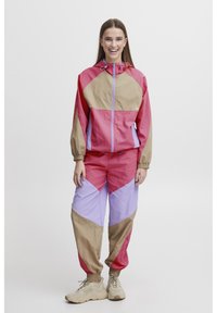 Rompevientos y pantalones de chándal coloridos hechos de tela ligera. Presenta patrones geométricos en rosa, púrpura y beige. Puños elásticos y capucha.