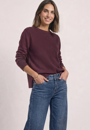 Femme souriante portant un pull bordeaux, un jean bleu, des boucles d'oreilles créoles dorées et un bracelet, debout les mains dans les poches devant un fond clair.