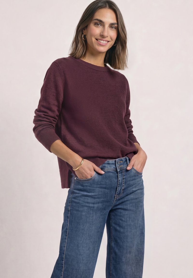 Femme souriante portant un pull bordeaux, un jean bleu, des boucles d'oreilles créoles dorées et un bracelet, debout les mains dans les poches devant un fond clair.