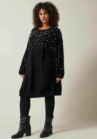 Abito maglione nero oversize con dettagli in perle, maniche lunghe e orlo a coste. Abbinato a jeans skinny neri e stivaletti.
