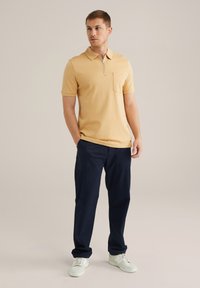 Polo cu mânecă scurtă în galben deschis, cu guler, detaliu cu fermoar și buzunar pe piept, purtat cu pantaloni închis la culoare și încălțăminte albă.