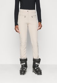 Beige skibroek met een slim fit, voorzien van twee ritszakken aan de voorkant en een gladde textuur, gedragen met zwarte skijschoenen.