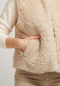 Gilet beige flou avec un extérieur texturé et duveteux, doté de poches latérales et d'une coupe décontractée, associé à un léger pull en dessous.