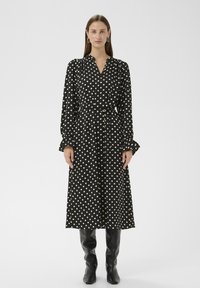 Schwarzes Kleid mit weißen Punkten, langen Ärmeln und einem taillierten Gürtel, mit Kragen und Rüschen an den Manschetten, kombiniert mit kniehohen schwarzen Stiefeln.