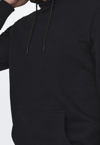 Sudadera negra con capucha fabricada en tela suave, que incluye un bolsillo frontal, capucha con cordón y puños acanalados. Textura lisa sin patrones visibles.