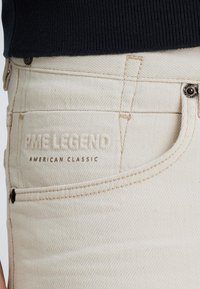 Jean en denim beige avec une texture subtile, comportant une poche cousue, un bouton métallique et un logo embossé portant la mention "PME Legend American Classic."