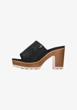 Sandalia plana negra tejida con un tacón grueso marrón. Presenta una parte superior texturizada y una entresuela en tono marrón claro contrastante. Incluye adornos decorativos.