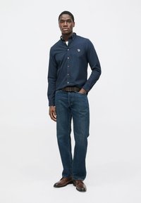 Tmavomodrá košeľa s dlhým rukávom a golierom na gombíky, kombinovaná s modrými džínsami a hnedými topánkami. Na hrudi je malý logotyp.