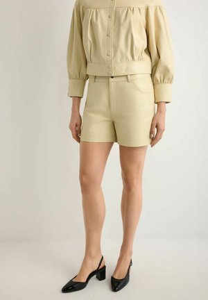 Kobieta ubrana w beżowy top na guziki z odkrytymi ramionami i dopasowane szorty oraz czarne sandały na słupku z paskiem z tyłu, stojąca na jasnym, jednolitym tle.